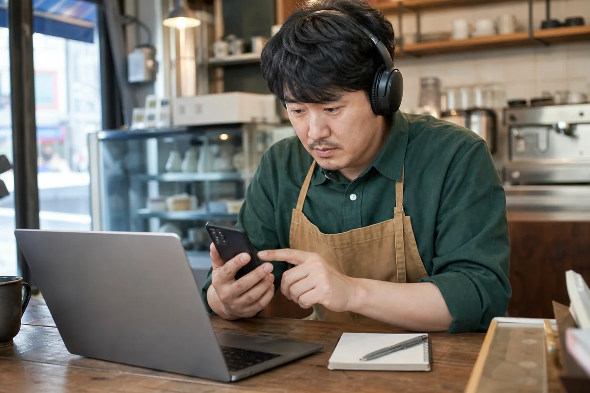 카페 운영자가 헤드폰을 끼고 노트북 옆에서 휴대전화의 오디오를 확인하는 실사풍 이미지